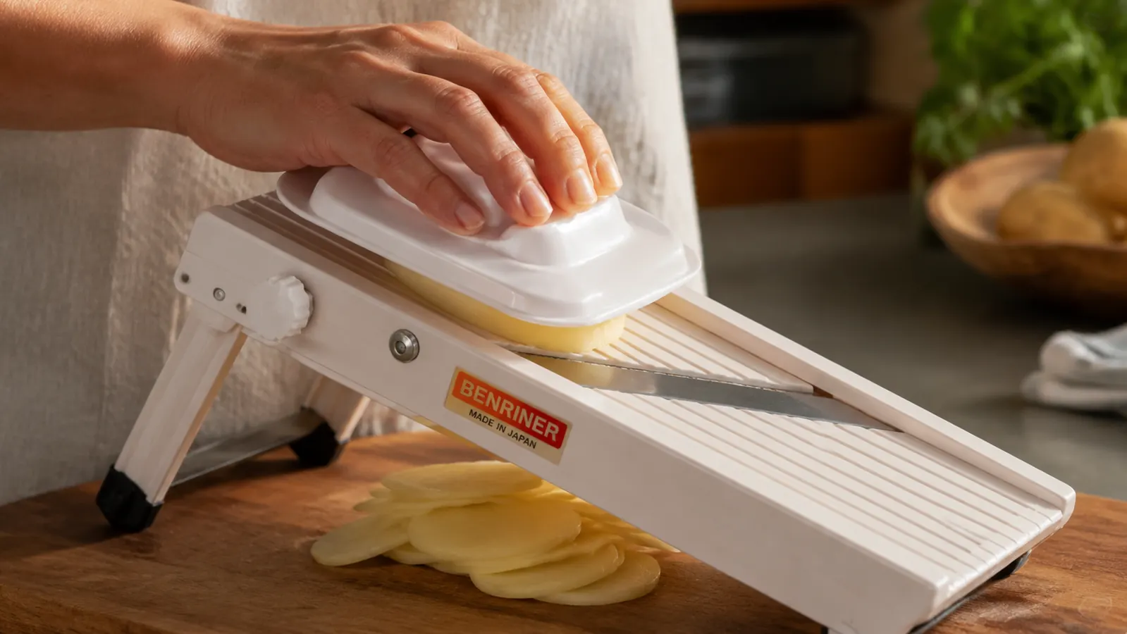 A hand sliding a potato across the Benriner mandoline blade with the hand guard in place, thin even slices falling onto a cutting board
