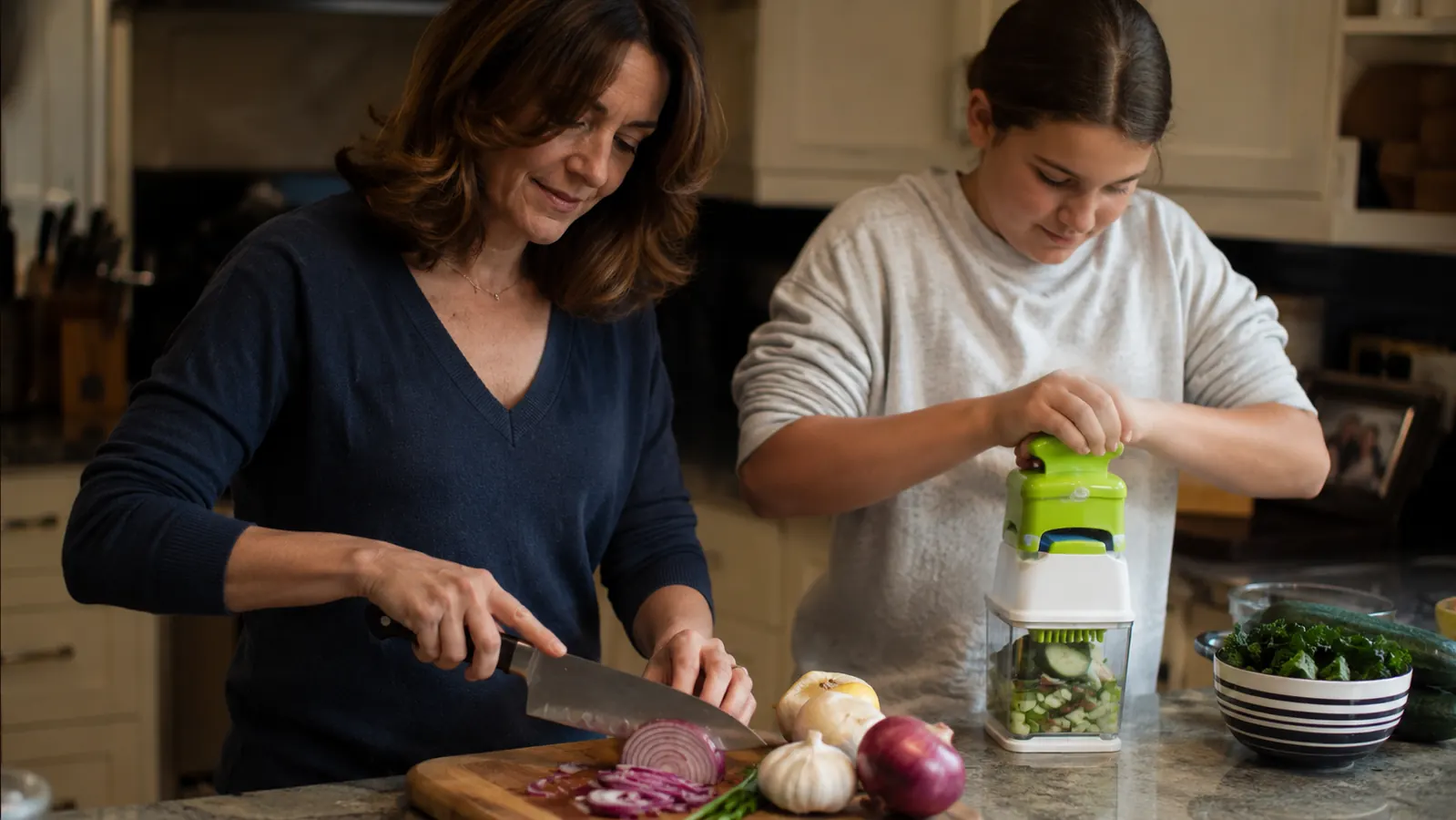 Two home cooks prepping vegetables side by side in a small kitchen, one using a chef's knife and one using a vegetable chopper